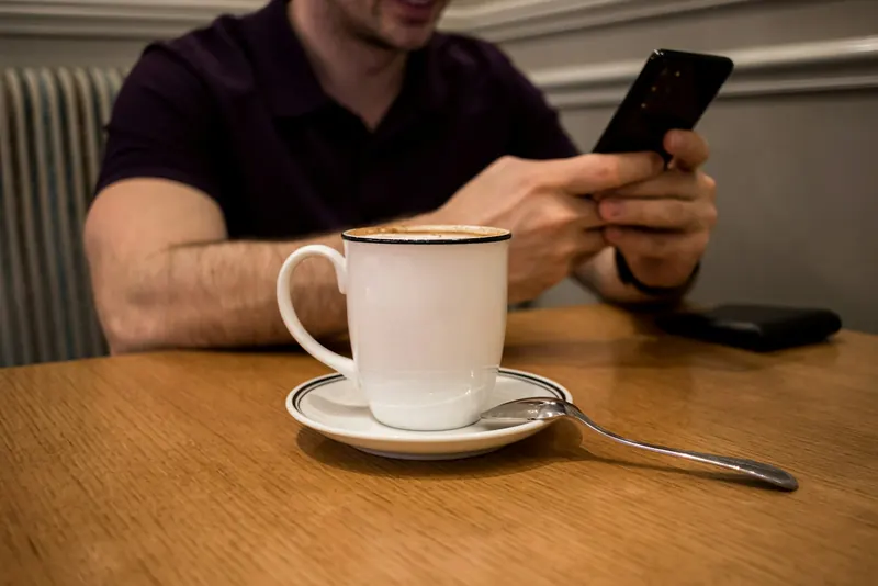 Un hombre lee en el celular, sentado a una mesa con una taza de café.